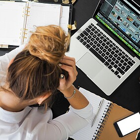 Das Foto zeigt eine Person, die gestresst wirkt und den Kopf in den Händen hält. Auf dem Schreibtisch befinden sich ein Laptop, Papiere und ein Stift. Hinweis: Dr. Schnutenhaus bietet Anti-Stress-Therapie an.
