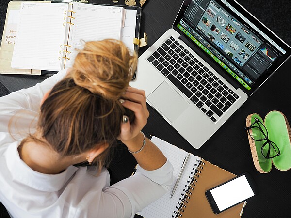 Anti-Stress durch Klopf- und Infusionstherapie Das Foto zeigt eine Person, die gestresst wirkt und den Kopf in den Händen hält. Auf dem Schreibtisch befinden sich ein Laptop, Papiere und ein Stift. Hinweis: Dr. Schnutenhaus bietet Anti-Stress-Therapie an.
