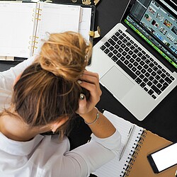 Das Foto zeigt eine Person, die gestresst wirkt und den Kopf in den Händen hält. Auf dem Schreibtisch befinden sich ein Laptop, Papiere und ein Stift. Hinweis: Dr. Schnutenhaus bietet Anti-Stress-Therapie an.